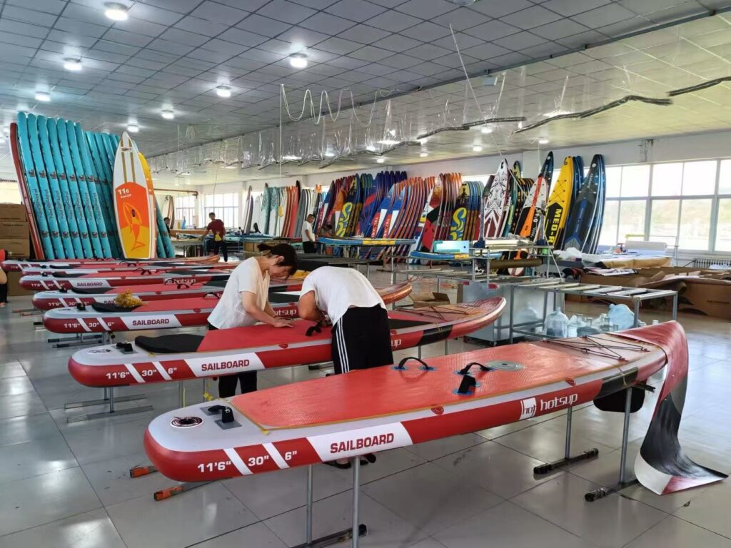 Workers assembling red inflatable SUP sailboards on a production line in a factory workshop, with a variety of finished stand-up paddleboards (SUPs) displayed in the background